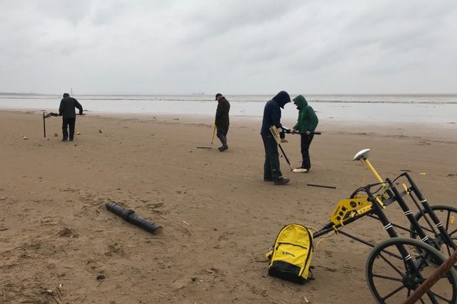 Four people on a beach, using equipment to find buried ordnance. In the foreground is a piece of scanning equipment on a wheel.